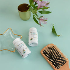 Zwei Flaschen SanaExpert Haar Forte Gold auf einer türkisen Oberfläche neben einer Pflanze und einem Holzhaarbürste.