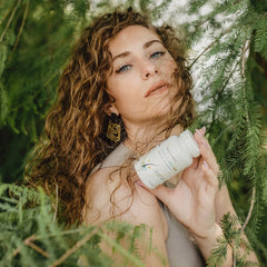 Eine Frau mit lockigen Haaren hält eine Flasche SanaExpert Haar Forte Gold, umgeben von grünen Pflanzen.