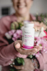 Lächelnde Frau hält eine Flasche SanaExpert Meno Forte, umgeben von blumiger Dekoration, freundliche und einladende Darstellung.