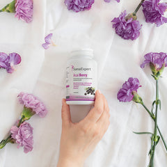 Hand hält eine Flasche SanaExpert Acai Berry Nahrungsergänzungsmittel, umgeben von violetten Blumen auf weißem Stoffhintergrund