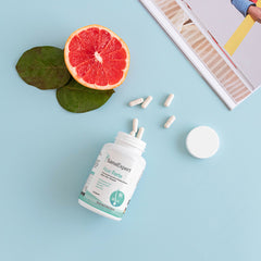 Eine geöffnete Flasche SanaExpert Haar Forte neben einer halbierten Grapefruit und Kapseln, arrangiert auf einem blauen Hintergrund.