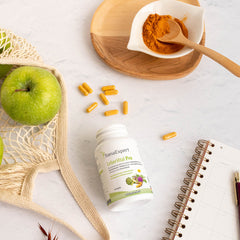 LeberVital Pro Dose auf einem hölzernen Tisch neben einem grünen Apfel, Kurkumapulver und Nahrungsergänzungskapseln, Notizbuch und Netzbeutel im Hintergrund.