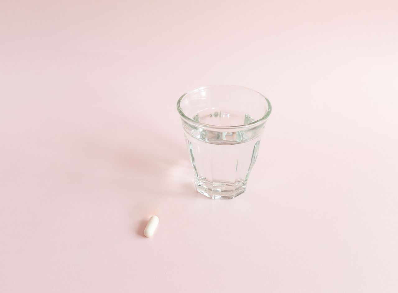 Minimalistisches Bild einer einzelnen weißen Kapsel neben einem klaren Wasserglas auf rosa Hintergrund, suggeriert eine einfache und reine Gesundheitsroutine.