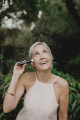 Eine lächelnde Frau mit grauen Haaren hält eine Flasche SanaExpert Collagen Beauty Elixir an ihr Ohr, als ob sie telefonieren würde, mit grüner Natur im Hintergrund.