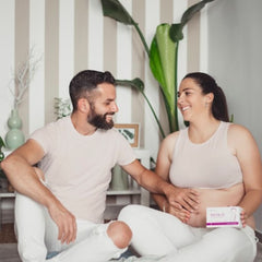 Lächelndes Paar sitzend im Wohnzimmer, die schwangere Frau hält eine Packung Natalis Pre, während der Mann seine Hand auf ihren Bauch legt.