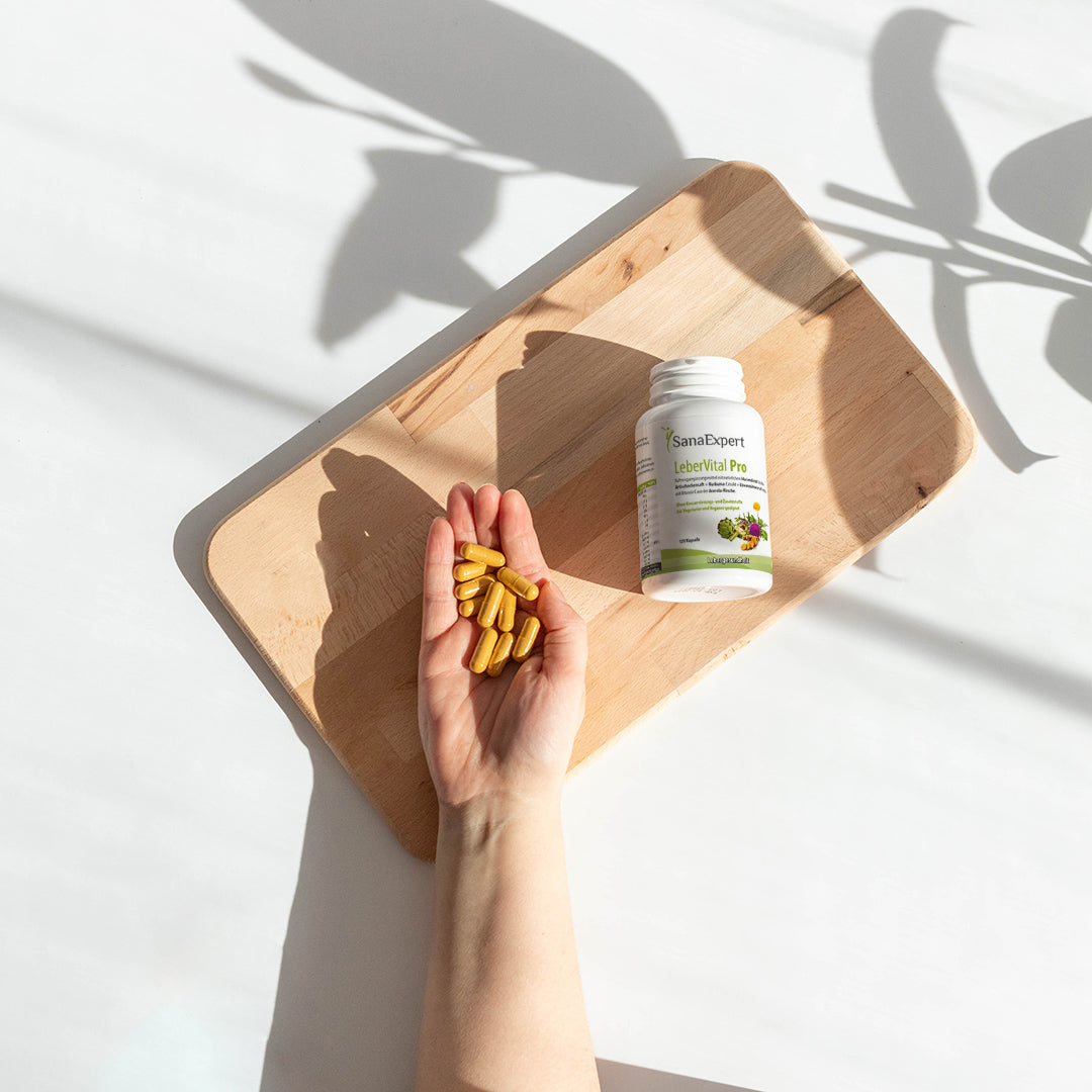 Eine Hand hält mehrere goldene Kapseln von LeberVital Pro über einem Holzbrett, mit einer Flasche des Produktes im Hintergrund, Schattenwurf von Pflanzen.