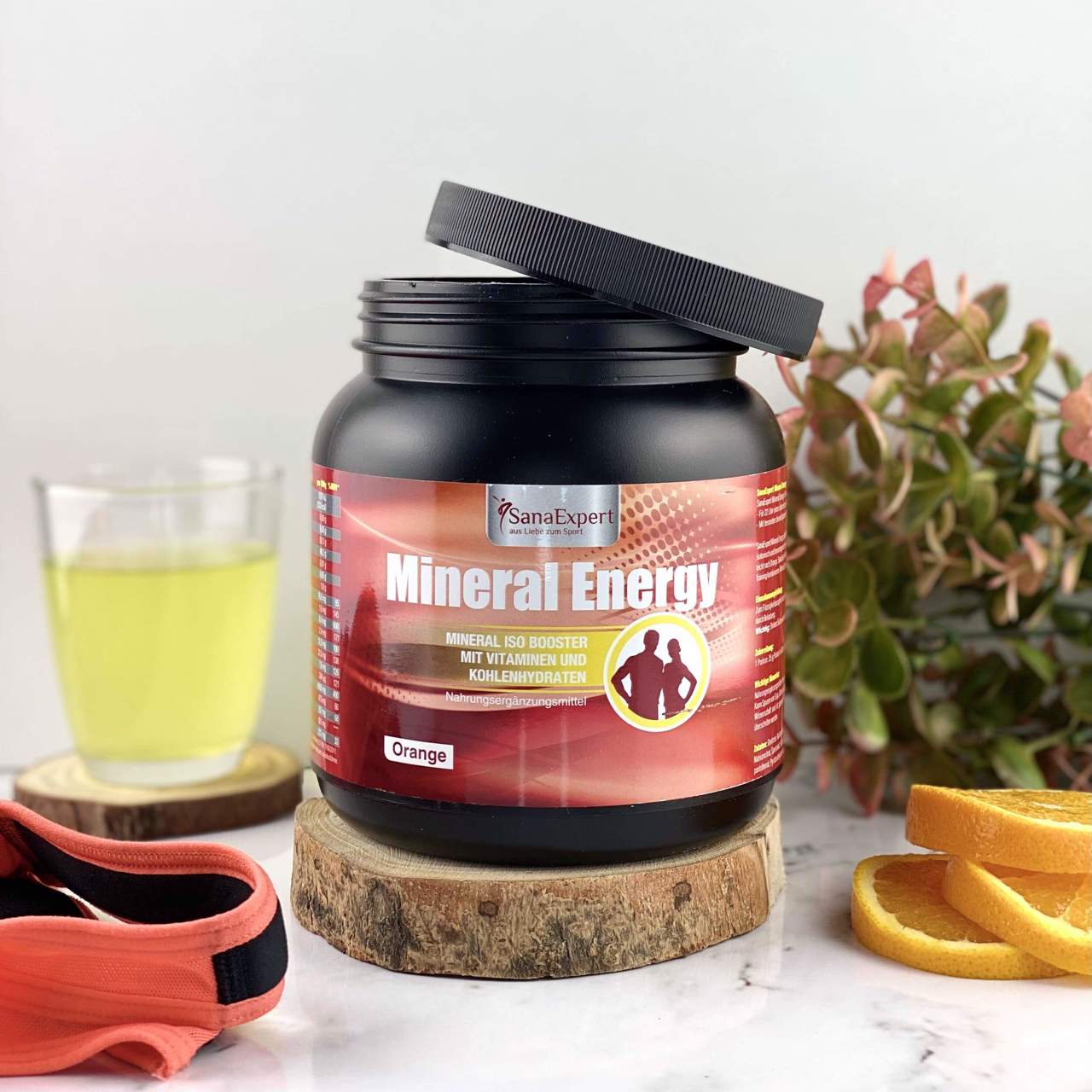 Nahaufnahme einer Mineral Energy Dose auf einem Holzstück, neben einem Fitnessband und Orangenscheiben, auf hellem Hintergrund.
