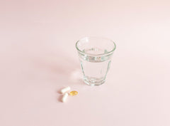 Minimalistische Darstellung einer Gesundheitsroutine mit einer weißen Kapsel und einer gelben Omega-3-Kapsel neben einem klaren Wasserglas auf rosa Hintergrund.